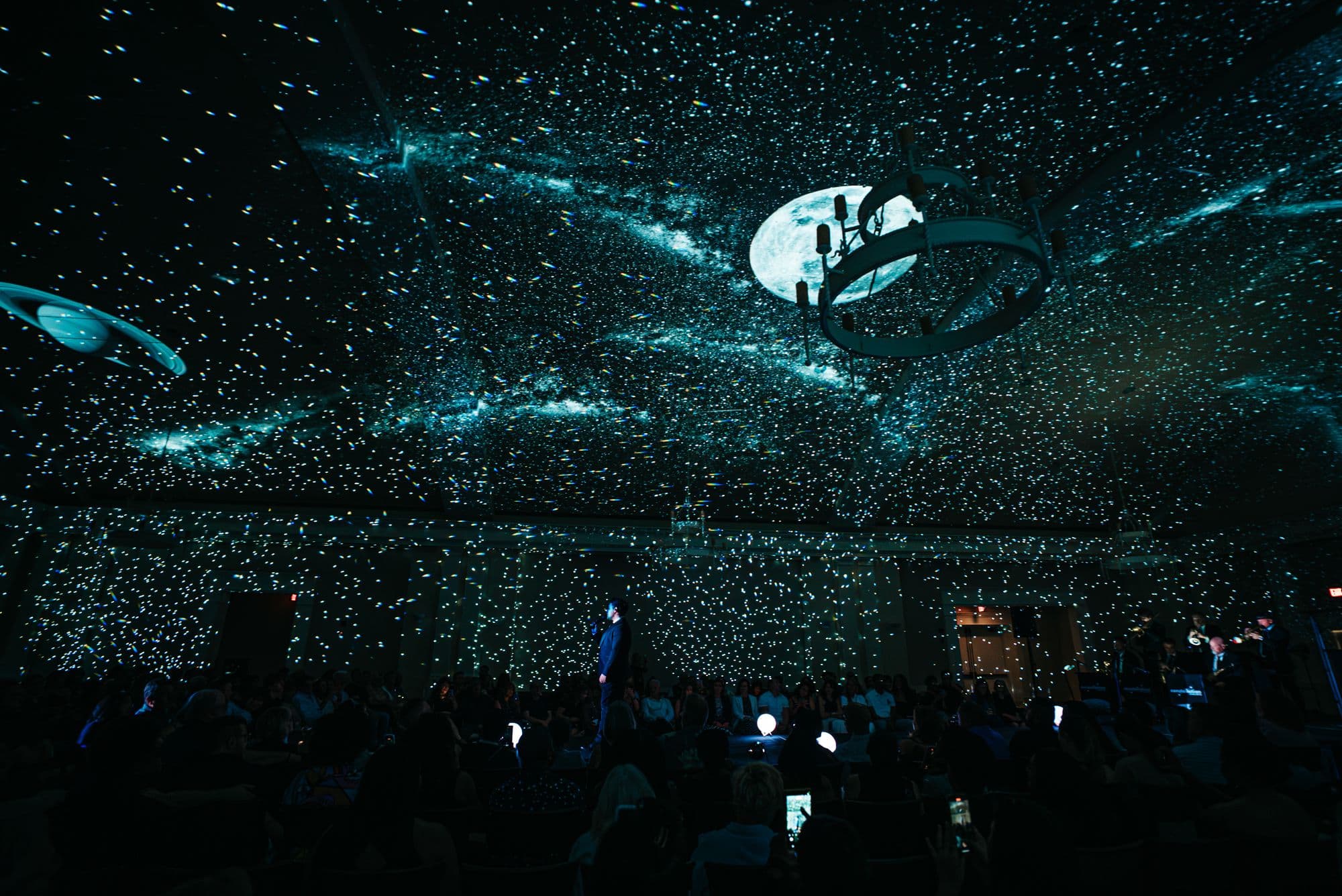A ballroom at a Constellation Concerts show: stars across every wall and the ceiling, the moon glowing behind a chandelier, the audience in silhouette